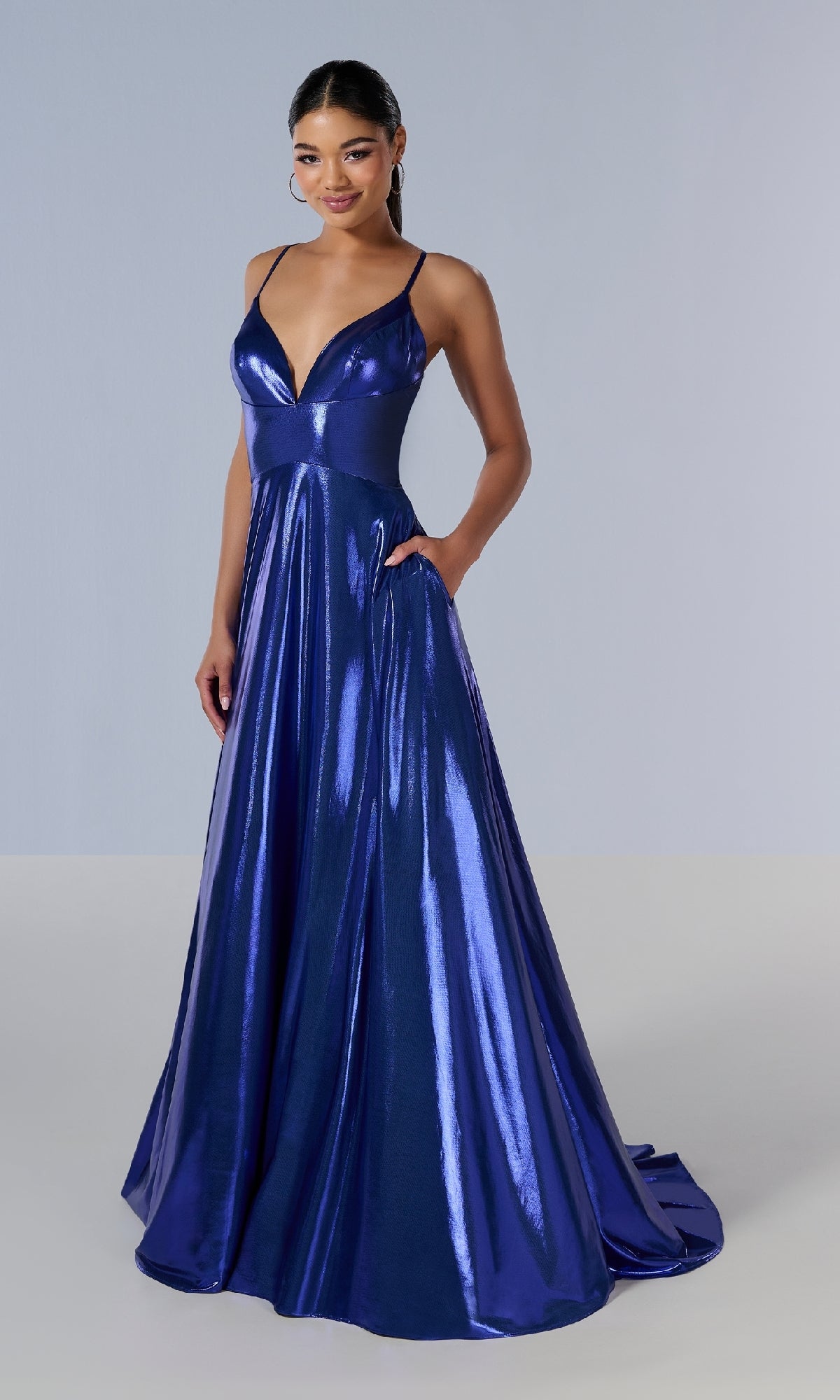 Woman models the Tiffany 16179 long prom dress, featuring a shiny metallic blue A-line silhouette with thin straps and a deep V neckline, standing against a plain background.